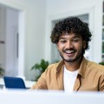 Indian student with curly hair studying sitting among books on shelves, man watching video course
